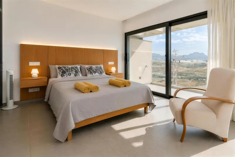 Dormitorio principal con cabecero de madera iluminado, sillón y puerta corredera al balcón
