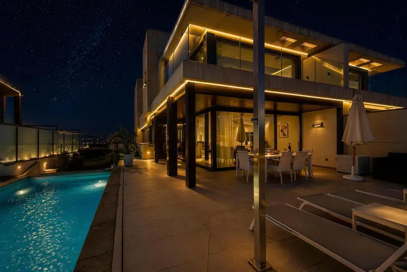 Fachada de la villa de noche, con luces cálidas, porche con comedor y piscina iluminada